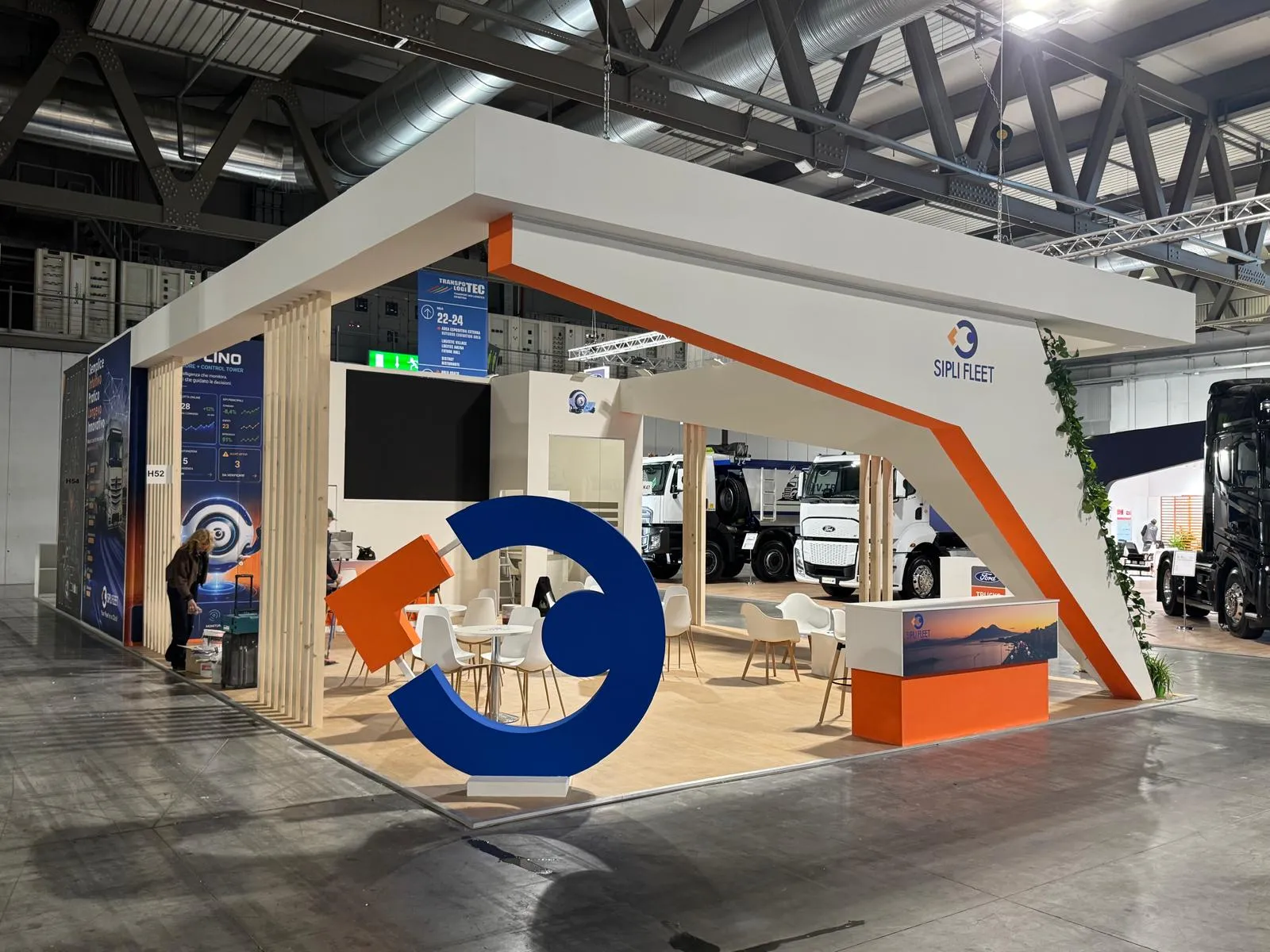 Stand SIPLI FLEET realizzato a Transpotec, vista d'angolo con colonna a V rivestita di edera e logo scultoreo blu e arancione a terra